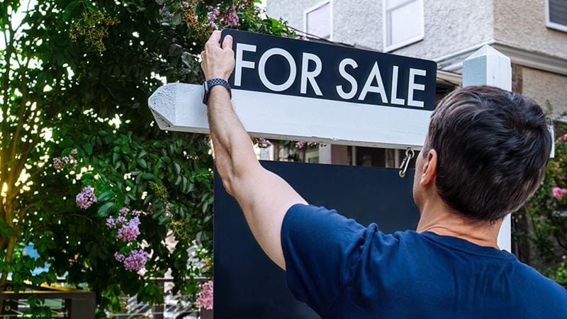 person putting for sale sign up in front of his house because home prices and interest rates may not decline any time soon.